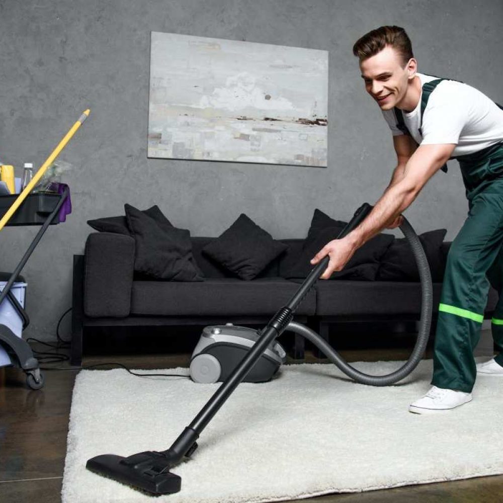 joven limpiador profesional sonriente limpiando alfombras con aspiradora
